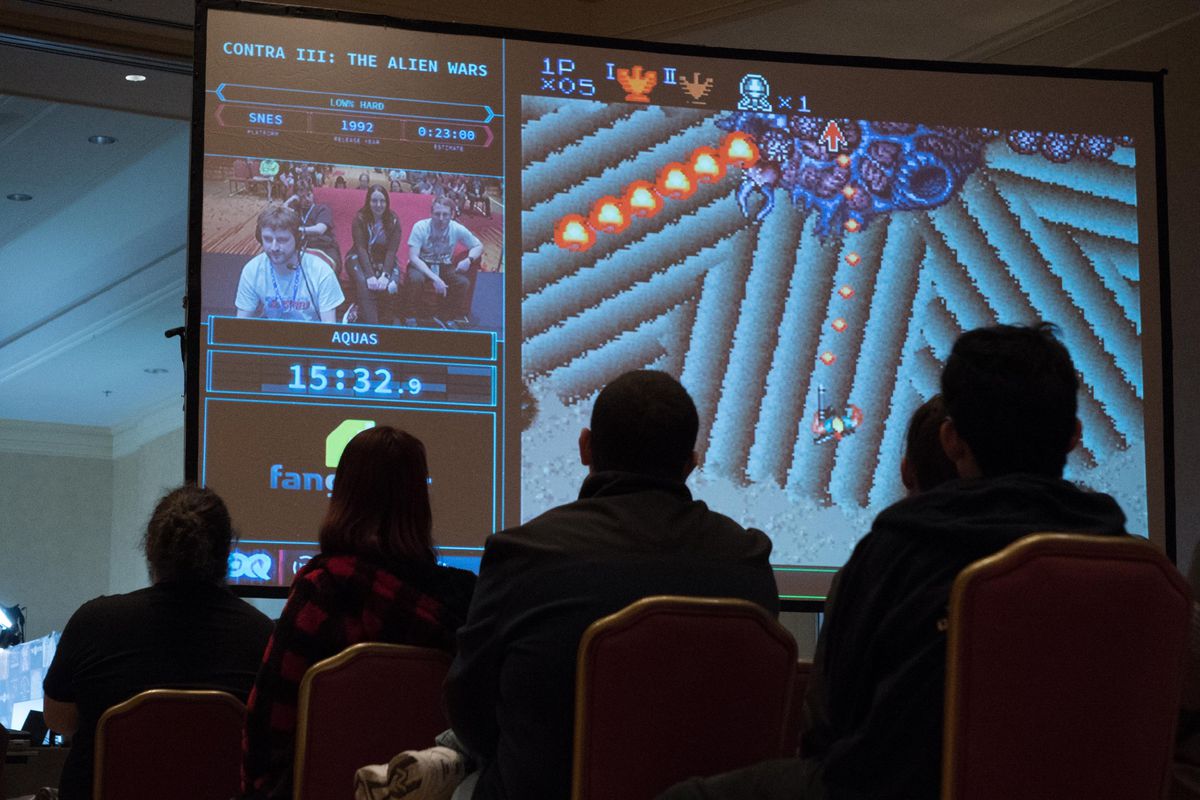 A photograph of the physical room where a speedrun of "Contra III: The Alien Wars" is taking place. The camera is at a low angle, and silhouettes of people sitting in the audience cover part of the large screen where the game is being projected.