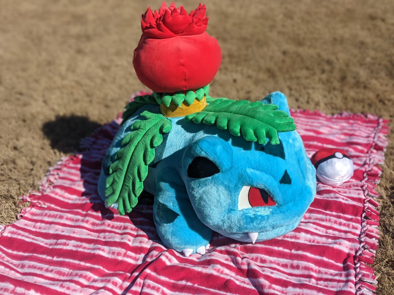 A stuffed animal of the Pokemon ivysaur, sitting on a beach towel in the sand on a sunny day, The plush is very large.