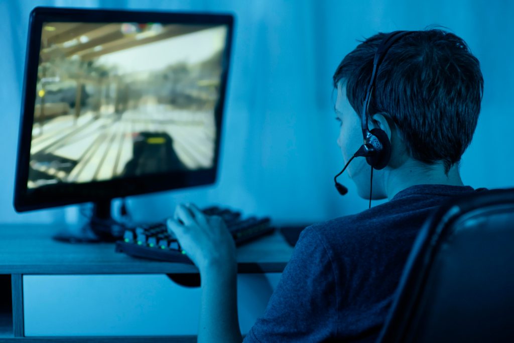 Young boy playing a video game on a computer. He could be streaming. He is wearing a headset with a microphone.