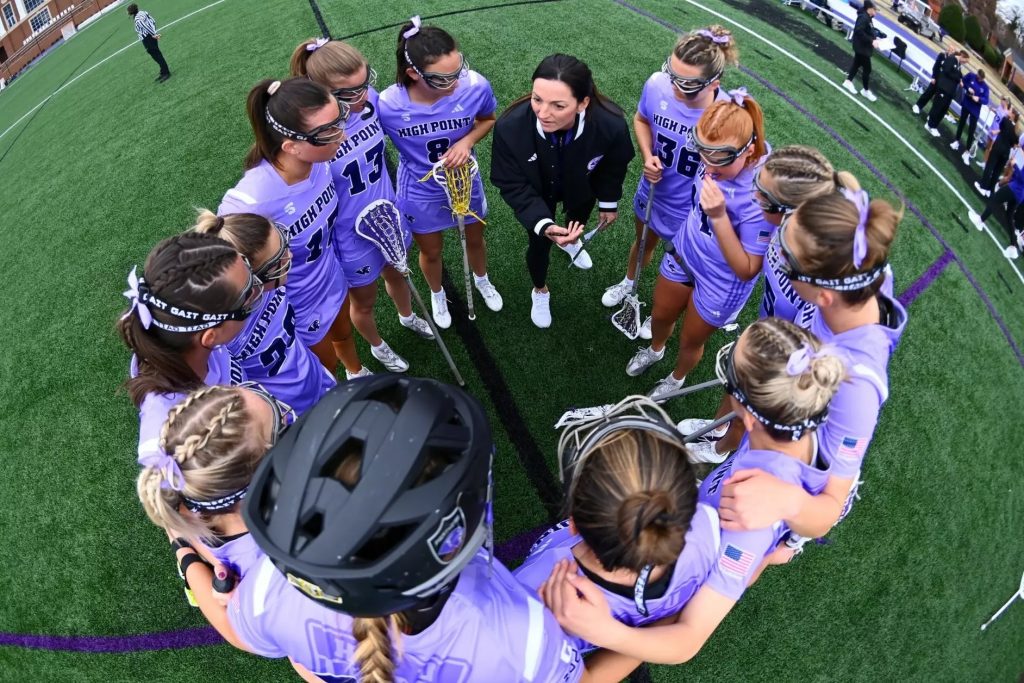 High Point University women’s lacrosse team in purple uniforms gathered in a tight huddle on the field, listening to their coach during a game.