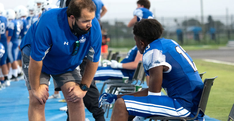 College football coach talking to a seated player in a blue uniform on the sideline during a game, with teammates and field visible in the background.
