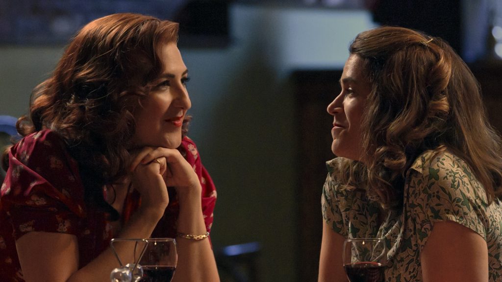 image shows two women sitting at a table. they are looking at each other lovingly. They both have their hair done in a forty's style.