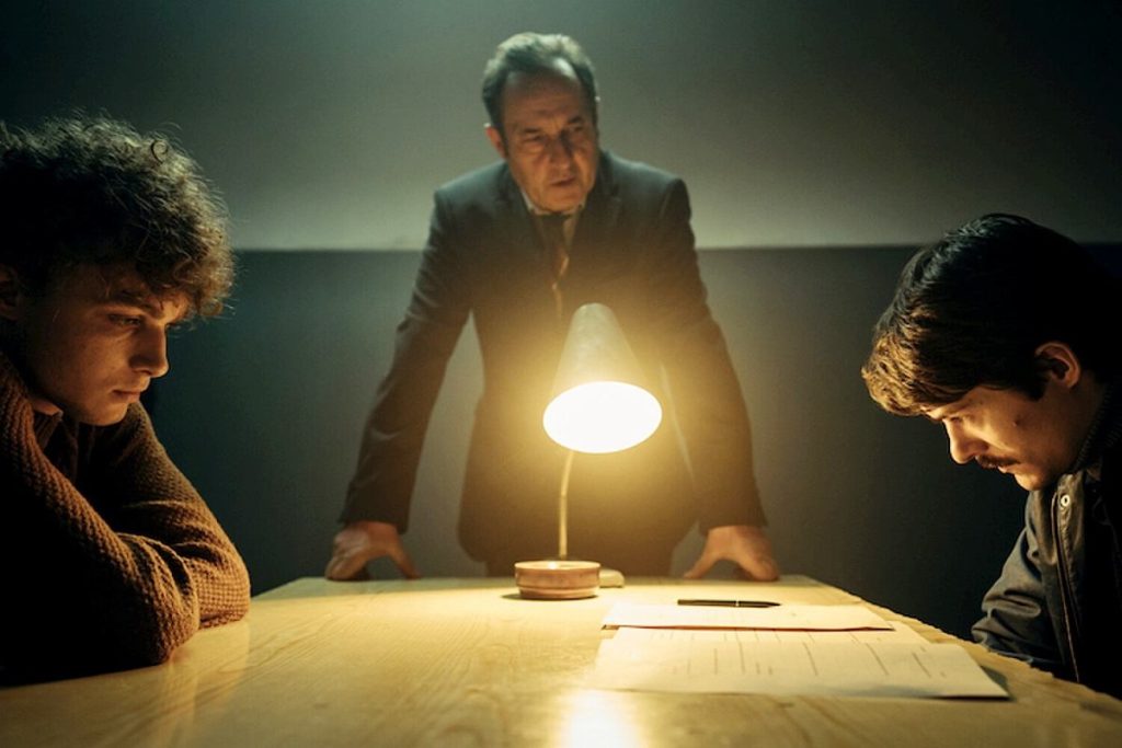 image shows three men. one is older and in the middle, he is interrogating the other two men who sit on either side of a table. there is also a bright light in the middle of the scene.