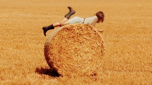 Image of a girl laying face down on a circular bale of hay, she has one foot up in the air as if she's kicking her legs back and forth. she's resting her weight on her elbows