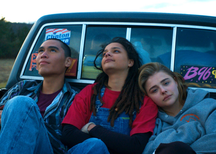 Image shows three teenagers sitting against the rearview window of a truck. The two to the left are looking at the sky and the one farthest to the right is looking at the camera. They all look hopeful.