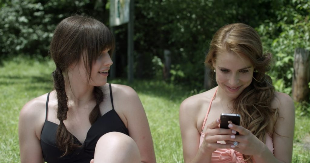 image is from Boy Meets Girl (2014). It shows two women sitting in bathing suits on a lawn. the woman on the left is looking at the woman to the right who is looking at her phone and smiling