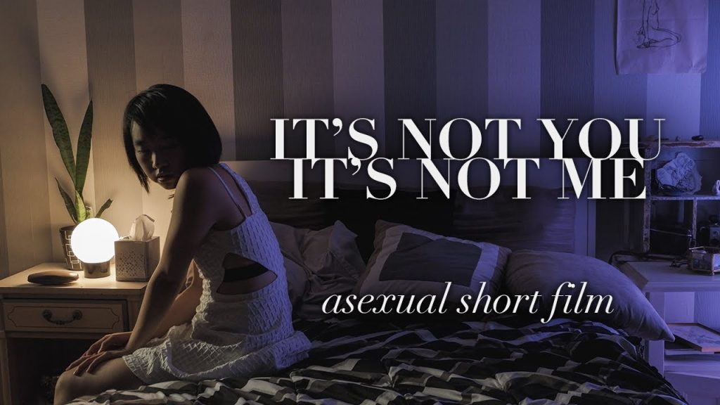 image from It's not You. It's Not Me. It shows a woman sitting on the edge of a bed facing towards the left-hand side of the screen. the right of the screen is mainly purple and the left is yellow. overtop of the image, text reads "It's Not You. It's Not Me" in bold text with "asexual short film" below it.