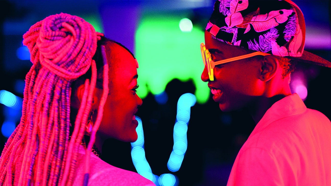 image is from Rafiki (2018). It shows two teenage girls looking at each other and smiling. Bright pink light engulfs them, there is bright purple and green light in the background.
