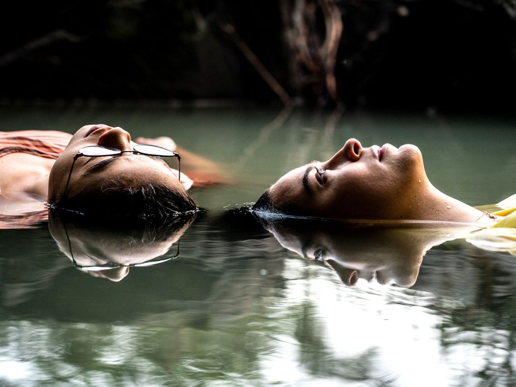 image from The Half Of It. its shows two teenage girls floating in water, their faces are reflected on the surface.